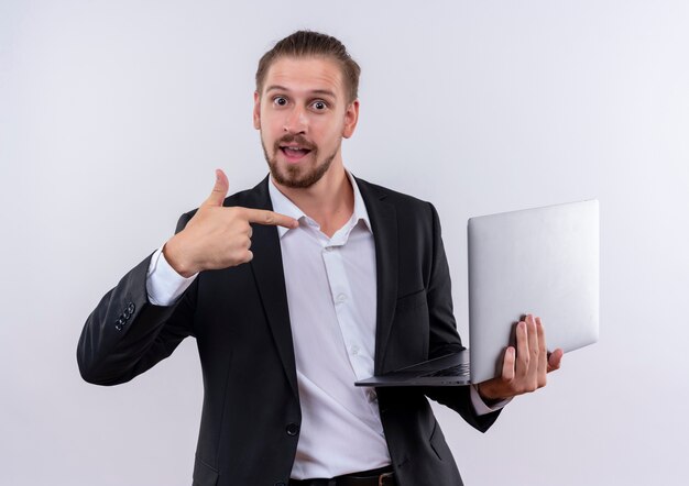 Apuesto hombre de negocios vestido con traje sosteniendo la computadora portátil apuntando con el dedo a ella sonriendo alegremente de pie sobre fondo blanco.