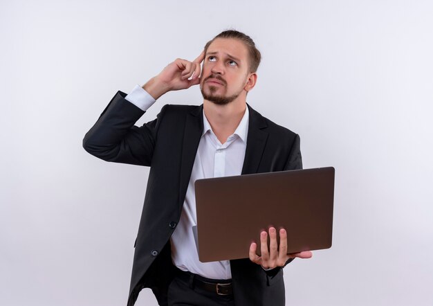 Apuesto hombre de negocios vestido con traje sosteniendo la computadora portátil mirando hacia arriba desconcertado parado sobre fondo blanco.