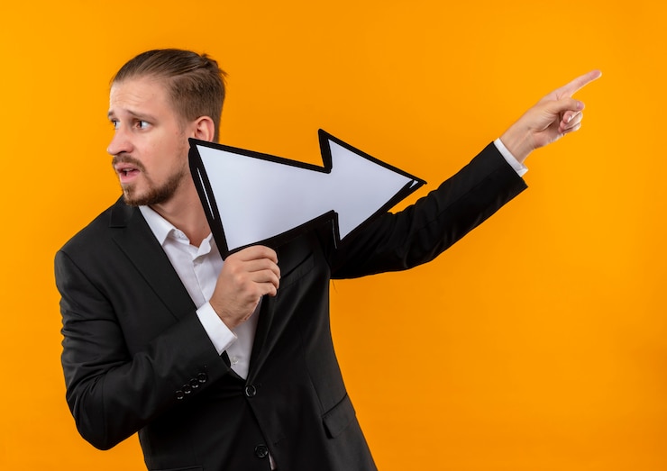 Foto gratuito apuesto hombre de negocios vestido con traje sosteniendo la flecha blanca apuntando con el dedo hacia el lado lookign confundido de pie sobre fondo naranja