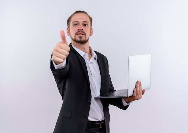 Apuesto hombre de negocios vestido con traje sosteniendo el ordenador portátil sonriendo alegremente mostrando los pulgares para arriba sobre fondo blanco.