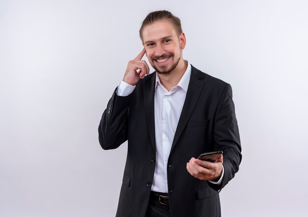 Apuesto hombre de negocios vestido con traje sosteniendo smartphone apuntando su templo con gran idea de pie sobre fondo blanco.