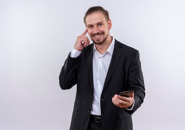 Apuesto hombre de negocios vestido con traje sosteniendo smartphone apuntando su templo con gran idea de pie sobre fondo blanco.