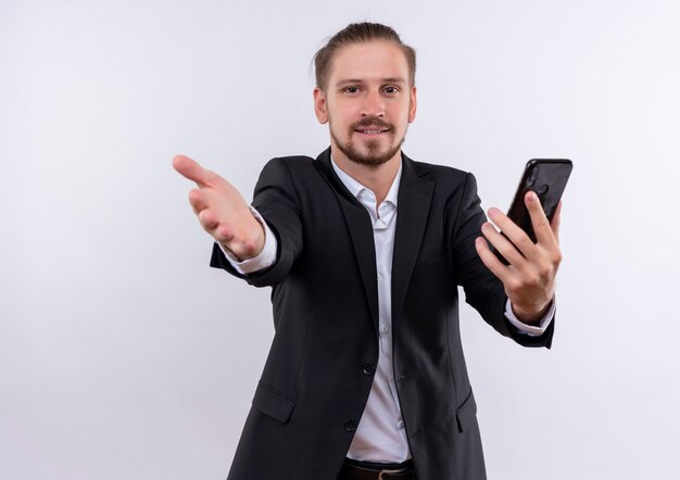 Apuesto hombre de negocios vestido con traje sosteniendo smartphone haciendo gesto de bienvenida con la mano sonriendo amable mirando a la cámara de pie sobre fondo blanco.