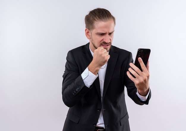 Apuesto hombre de negocios vestido con traje sosteniendo smartphone mirando a la pantalla mostrando puño con cara seria de pie sobre fondo blanco.