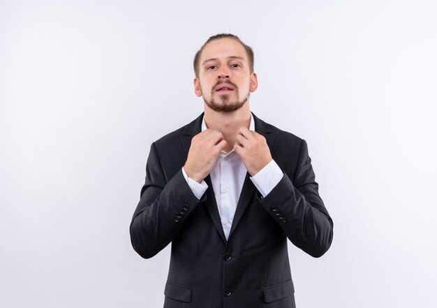 Apuesto hombre de negocios vestido con traje tocando su cuello mirando confiado sobre fondo blanco.