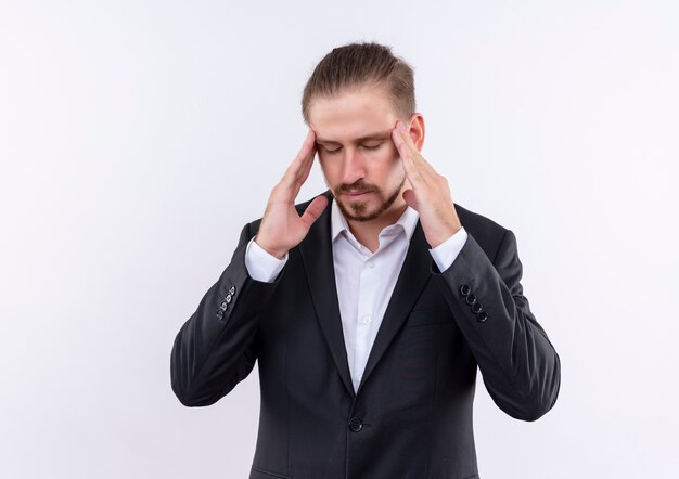 Apuesto hombre de negocios vestido con traje tocando sus sienes con aspecto cansado y con exceso de trabajo con dolor de cabeza de pie sobre fondo blanco.