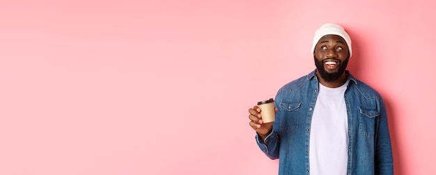 Apuesto hombre negro moderno bebiendo café para llevar sonriendo y mirando satisfecho a la cámara de pie