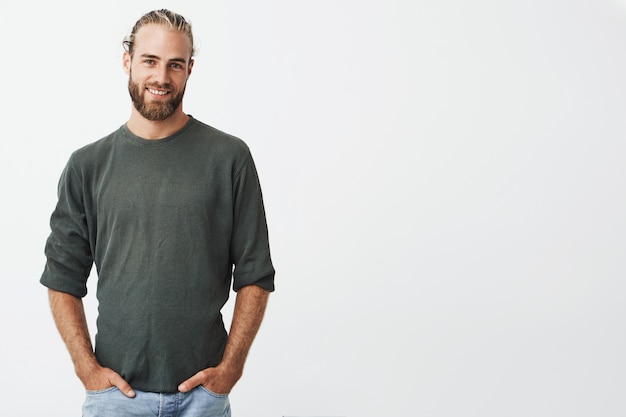 Apuesto hombre nórdico con barba y elegante peinado en camisa gris y jeans sonriendo, mantiene las manos en los bolsillos.