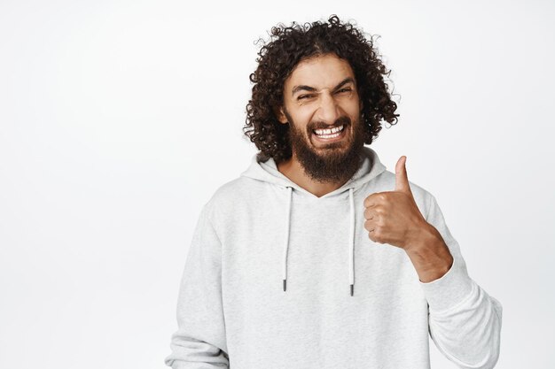 Apuesto hombre de Oriente Medio con barba mostrando los pulgares hacia arriba y sonriendo alabanza y cumplido algo bien parecido satisfecho fondo blanco