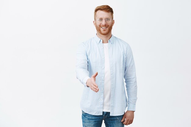 Apuesto hombre pelirrojo amable y encantador con cerdas en gafas y camisa, tirando de las manos hacia en gesto de apretón de manos y sonriendo felizmente