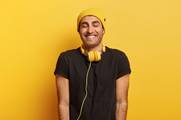 Apuesto hombre positivo con auriculares en el cuello, sonríe ampliamente y cierra los ojos de placer, usa sombrero y camiseta negra