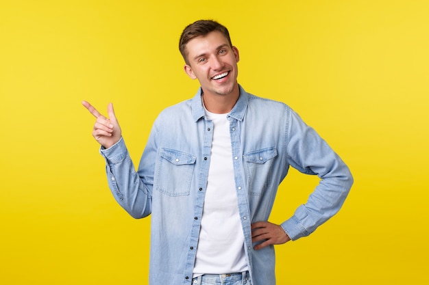 Apuesto hombre rubio adulto carismático con una sonrisa blanca perfecta, presentar un nuevo producto, señalar con el dedo en la esquina superior izquierda, demostrar un banner publicitario, fondo amarillo de pie