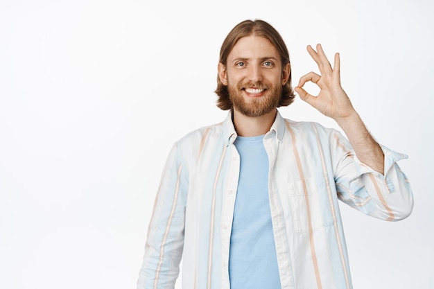 Un apuesto hombre rubio con barba que muestra un buen signo, de pie con una camisa blanca en el fondo del estudio