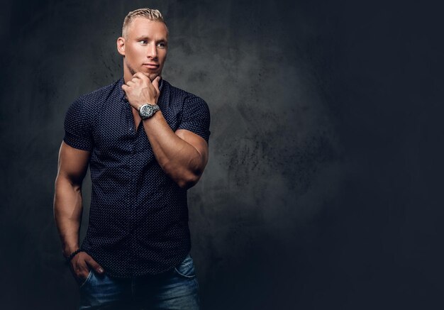 Un apuesto hombre rubio con una camisa azul oscuro posando sobre un fondo gris en un estudio.