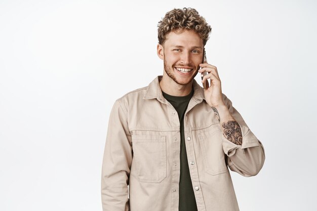 Apuesto hombre rubio sonriente hablando por teléfono móvil usando un teléfono inteligente haciendo una llamada de pie contra el fondo blanco