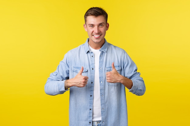 Apuesto hombre rubio sonriente que muestra el pulgar hacia arriba, anima a la persona, apoyándote. Chico complacido recomendando producto, dejar comentarios positivos, dar me gusta y aprobar el servicio, fondo amarillo.