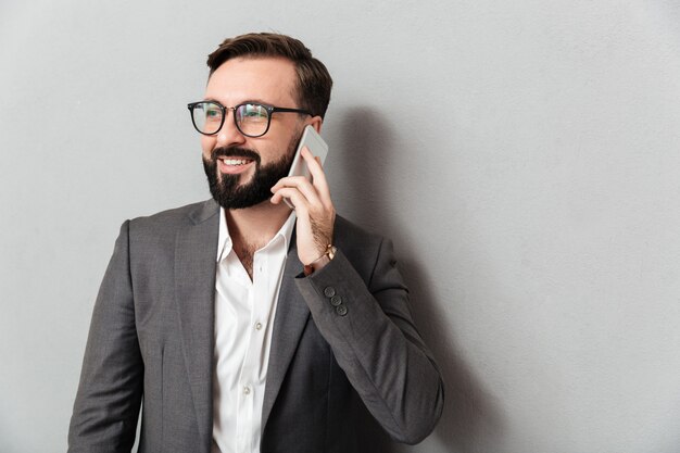 Apuesto hombre serio en ropa formal teniendo conversación móvil con smartphone aislado sobre gris