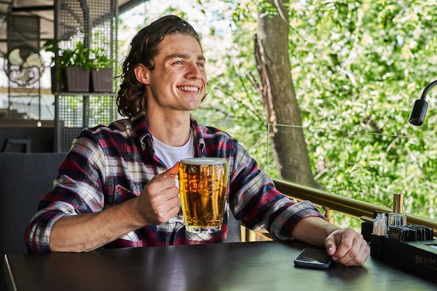 Apuesto hombre sonriente bebiendo cerveza en la cafetería de la terraza de verano.