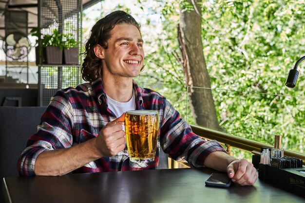 Apuesto hombre sonriente bebiendo cerveza en la cafetería de la terraza de verano.