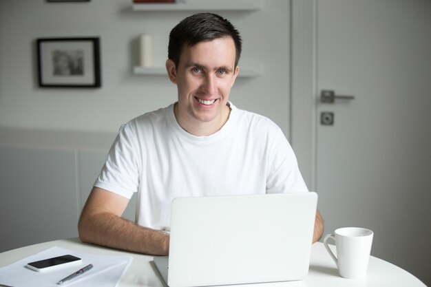 Apuesto hombre sonriente en el escritorio blanco trabajando con ordenador portátil