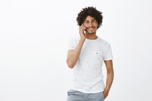 Apuesto hombre sonriente hablando por teléfono, casualmente de pie sobre la pared blanca