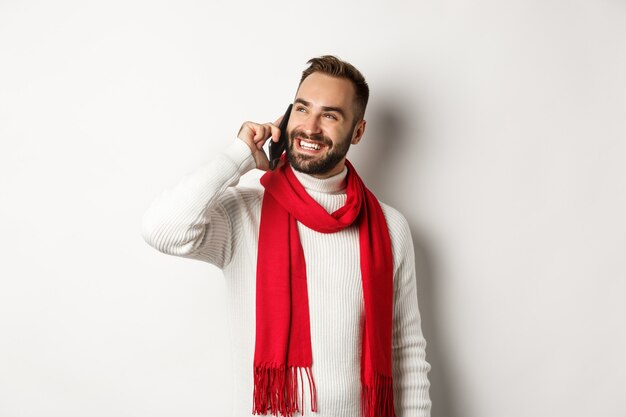 Apuesto hombre sonriente hablando por teléfono, mirando satisfecho, de pie en un suéter y bufanda de invierno, fondo blanco.