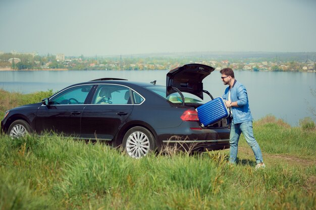 Apuesto hombre sonriente en jeans, chaqueta y gafas de sol yendo de vacaciones, cargando su maleta en el maletero del automóvil en el lado del río.