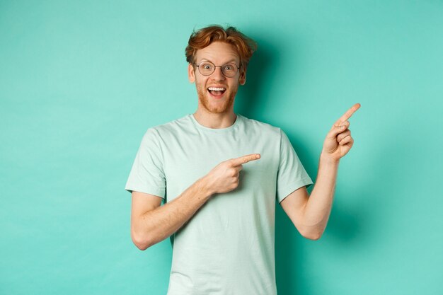 Apuesto hombre sonriente con pelo rojo y barba, mirando divertido y apuntando a la esquina superior derecha, mostrando la oferta promocional, de pie sobre fondo de menta