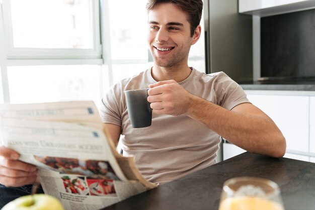 Apuesto hombre sonriente con una taza de té leyendo el periódico en la cocina