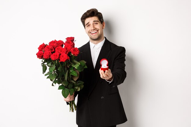 Apuesto hombre sonriente en traje negro, sosteniendo rosas y anillo de compromiso, haciendo una propuesta para casarse con él, de pie contra el fondo blanco.