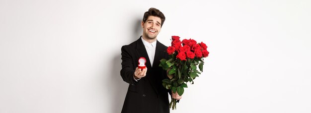 Un apuesto hombre sonriente con traje negro sosteniendo rosas y un anillo de compromiso haciendo una propuesta para casarse con él