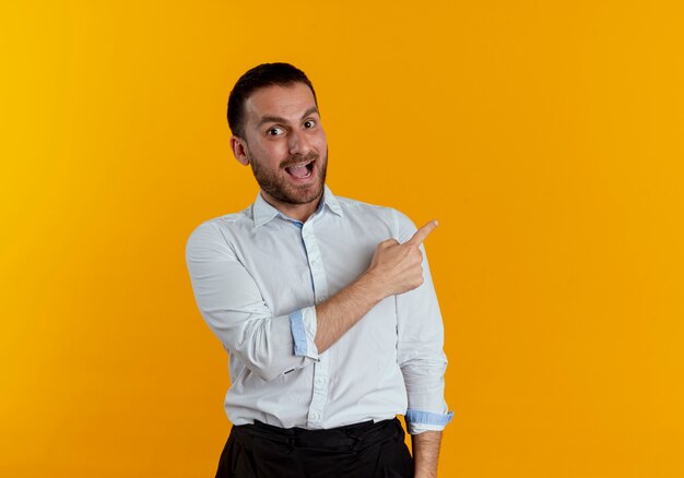 Apuesto hombre sorprendido apunta al lado mirando aislado en la pared naranja