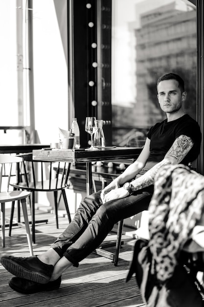 Apuesto hombre tatuado en una terraza de verano en un café de la ciudad está bebiendo vino. Calle Cafe.