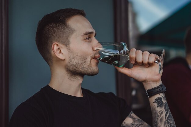 Apuesto hombre tatuado en una terraza de verano en un café de la ciudad está bebiendo vino. Calle Cafe.