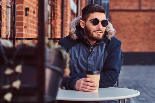 Un apuesto joven con un abrigo de invierno sentado con café en la cafetería al aire libre.