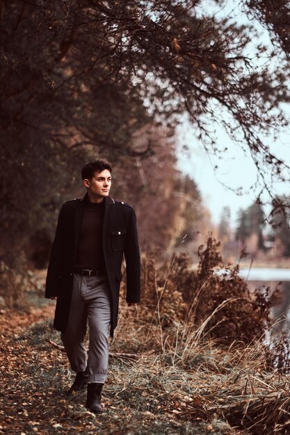 Un apuesto joven con un abrigo negro caminando cerca de un lago en el bosque de otoño.