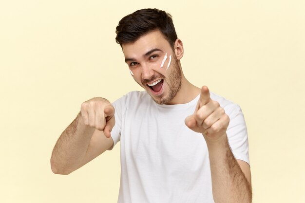 Apuesto joven sin afeitar en camiseta blanca sonriendo y señalando con el dedo índice al frente