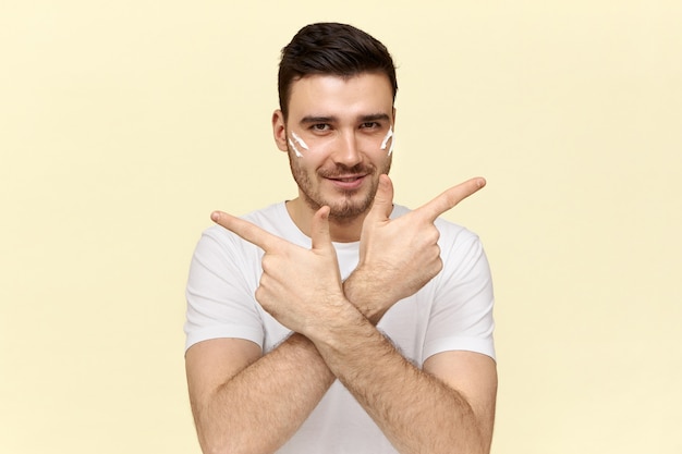 Apuesto joven sin afeitar en camiseta blanca sonriendo y señalando con el dedo índice a los lados