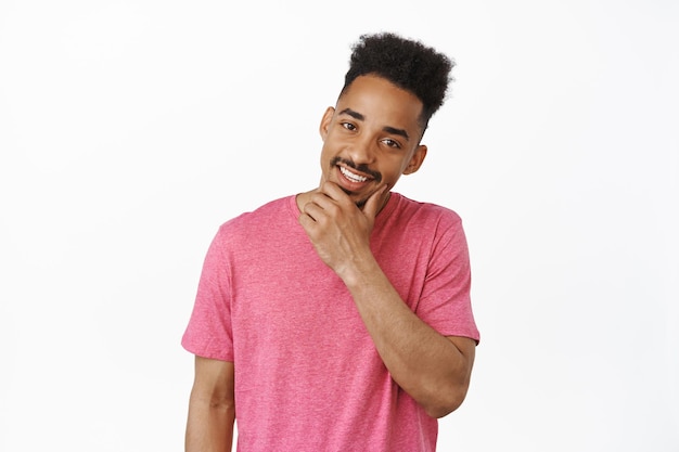 Un apuesto joven afroamericano con un elegante corte de pelo, tocándose la barba y sonriendo los dientes blancos, de pie con una camiseta rosa informal sobre fondo blanco