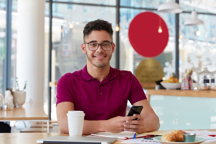 Foto gratuito apuesto joven alegre busca trabajo, navega por la página web en un teléfono celular moderno, verifica información en internet, se sienta en la cafetería, disfruta de un postre dulce y una bebida fresca