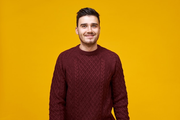 Apuesto joven alegre con elegante corte de pelo y sonrisa con hoyuelos posando aislado contra la pared amarilla en blanco, vestido con un acogedor suéter marrón, con mirada de confianza