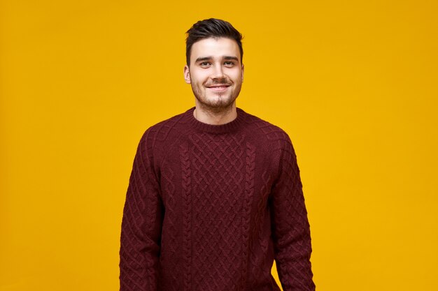 Apuesto joven alegre con elegante corte de pelo y sonrisa con hoyuelos posando aislado contra la pared amarilla en blanco, vestido con un acogedor suéter marrón, con mirada de confianza