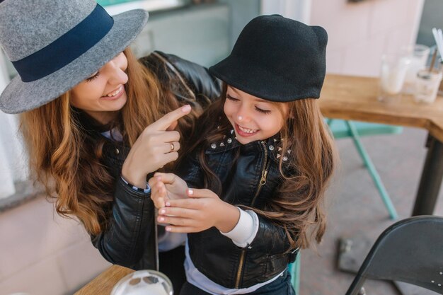 Apuesto joven apuntando con el dedo a su pequeña hija sonriente y riendo. Retrato de mamá alegre y su niño con estilo feliz descansando juntos en la cafetería al aire libre después de ir de compras en un día soleado