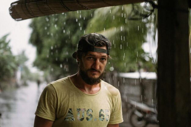 Un apuesto joven barbudo con una gorra bajo la lluvia tropical.