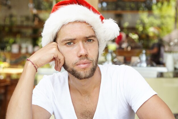 Apuesto joven barbudo con sombrero de Santa Claus mirando a la cámara, con expresión triste y aburrida en su rostro sintiendo nostalgia