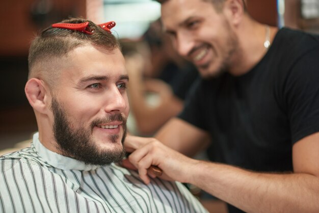 Apuesto joven barbudo sonriendo mirando a otro lado mientras el peluquero profesional le da un copyspace de corte de pelo.