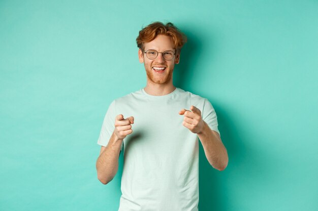 Apuesto joven con cabello pelirrojo, con gafas y camiseta, apuntando con el dedo a la cámara y sonriendo, eligiéndote, felicitándote o alabando, de pie sobre fondo de menta