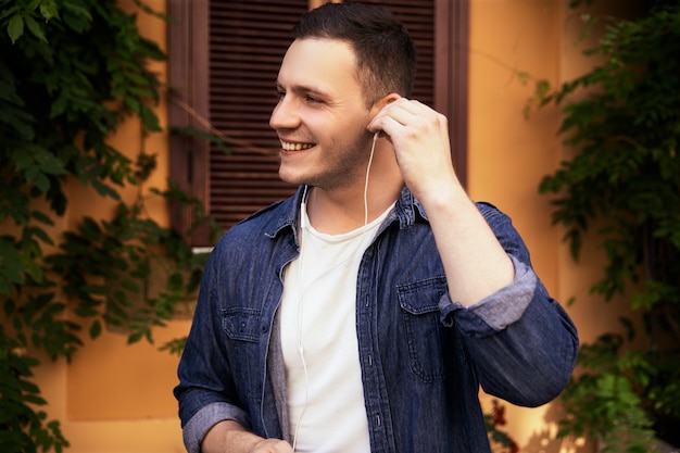 Apuesto joven en una camisa de jeans está escuchando música en los auriculares al aire libre