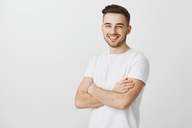 Apuesto joven en camiseta blanca, pecho de brazos cruzados y sonriendo complacido