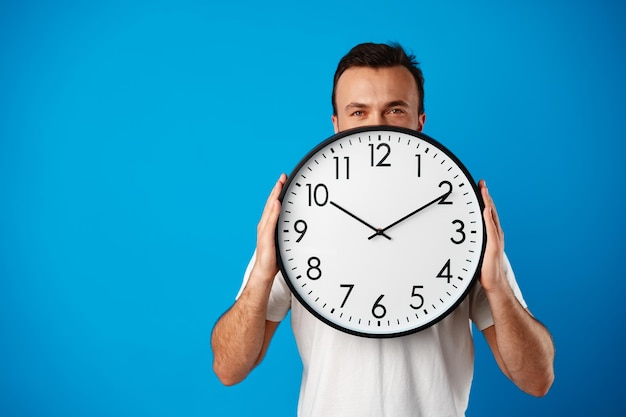 Apuesto joven en camiseta blanca posando con reloj sobre fondo azul.